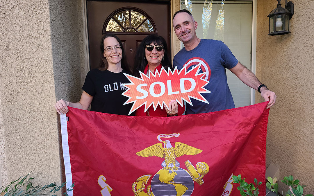 Real estate agent Maria Casas, veteran Sean Amman, and wife holding a sold sign and Marine Corps flag in front of their new house.