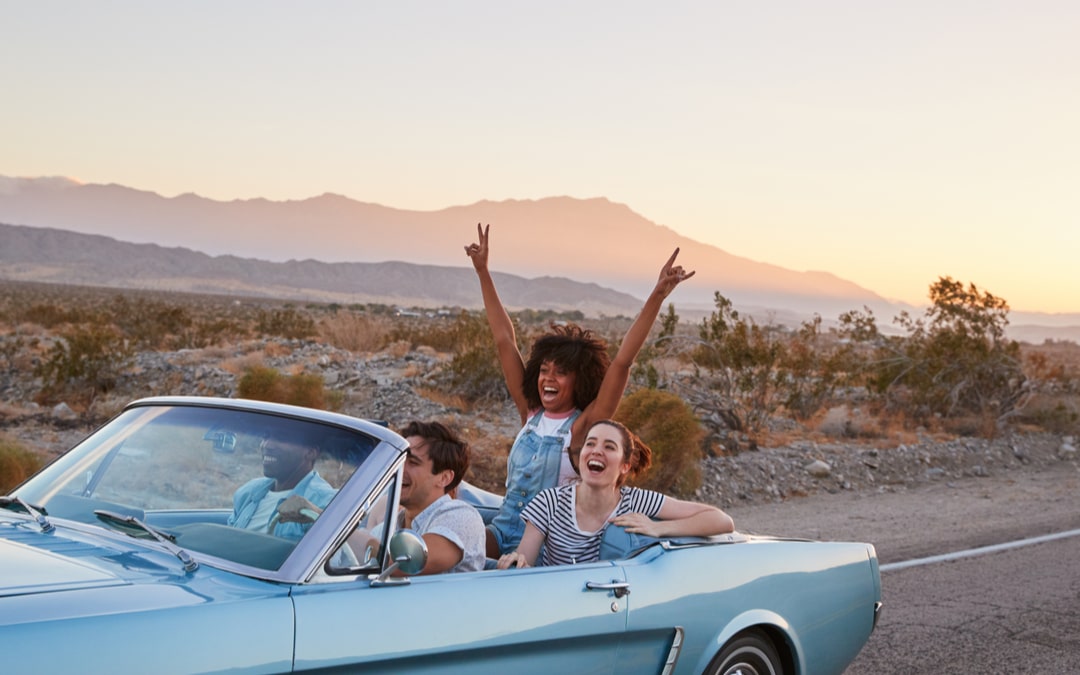 Three people in convertible top blue car driving in the desert at sunset